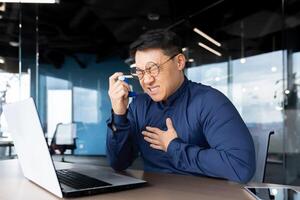 Asthma attack at work, asian businessman having difficulty breathing, man using inhaler and medicine to ease breathing, worker inside office with laptop at work. photo
