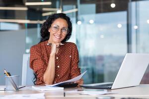 Portrait of happy and successful hispanic woman, businesswoman smiling and looking at camera holding contracts and invoices, working inside office with laptop on paper work. photo
