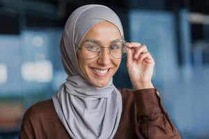Close-up photo of a young beautiful woman in a hijab and glasses looking at the camera and smiling, a student close-up studying inside a modern university.