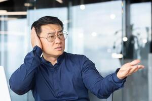 Excessive noise in the office, a businessman tries to be quiet, a man tries to concentrate on work, is disturbed by colleagues in the office, an Asian uses a laptop at work, nervous and angry. photo