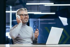 Senior smiling gray-haired man sitting at desk in office wearing headphones in front of laptop. Talks via call, listens to music, webinars, studies, teaches, rests. photo