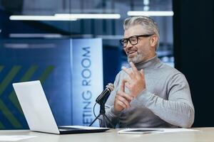 Senior gray-haired man sitting at a desk in an office with a microphone and laptop. Talks on a call. Conducts interviews, business meetings, webinars, online meetings. Shows, counts on fingers. photo