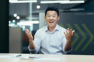 Portrait of a young happy Asian man sitting at an office desk in front of a camera and talking on a call. Gesturing with his hands, smiling at the camera. photo
