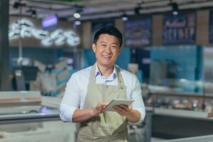 Portrait of a handsome young Asian man, an employee of a store, supermarket. Standing in an apron, holding a notepad, looking at the camera, smiling photo