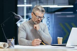 Close-up photo. Concentrated senior gray-haired man architect, engineer, designer sitting in office at table with laptop, working with drawings.plans. Makes notes, paperwork. photo