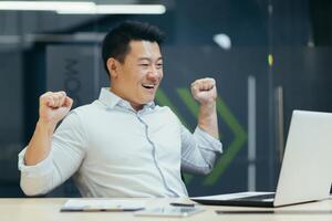 Asian businessman works in a modern office, rejoices at the end of the working day, and is satisfied with the work done, the man looks at the laptop screen and holds his hands up. photo