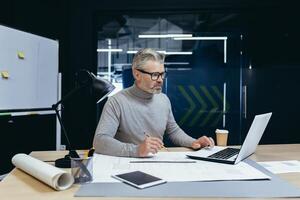 Senior gray-haired man architect, engineer, designer sits in the office at the table, works on a laptop, drawings. Makes notes. photo