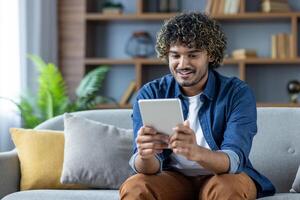 Young man sitting on sofa in living room at home with tablet computer in hands, smiling happily, using online app, browsing social media photo