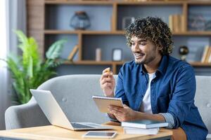 A young man is engaged in online learning, sitting on a comfortable sofa with his laptop and a notebook in hand. He appears focused and enthusiastic, using digital tools for his studies. photo