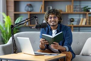 Portrait of a book author blogger, a man is recording an audiobook sitting at home on a sofa in the living room, looking at camera with a book in hands, using a laptop and a professional microphone photo