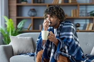 Sick man with a cold sits at home on the couch, covered with a blanket, and with a napkin in his hand. A man with a cold with a hot drink in his hands has a runny nose photo