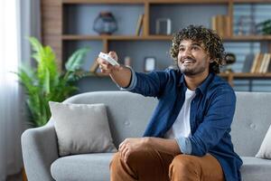 Joyful man turns on the air conditioner, sits with the remote control in his hands from the smart home system, on the sofa in the living room at home photo