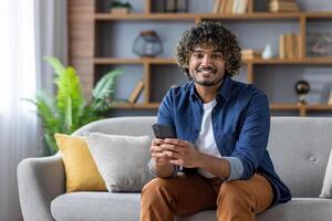 Young man with phone in hands smiling and looking at camera, using app on smartphone, sitting on sofa in living room of apartment at home photo