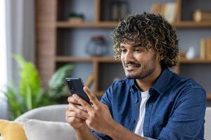 A young man is smiling while holding a smartphone. He is casually dressed, creating a relaxed and comfortable atmosphere. The focus is on his engagement with the phone. photo