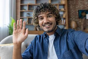 A man looks into the phone camera, happily waves his hand, takes a selfie photo and talks to colleagues and friends remotely using a smartphone app for chats