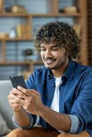 Young man at home using an app on his phone, sitting on a sofa in the living room holding a phone in his hands. Typing a text message smiling and browsing social networks, internet pages. photo