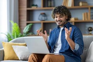 Joyful man sitting on sofa at home, using laptop for call, remote online communication, conference meeting. Businessman working from home in living room. Satisfied student watching course photo