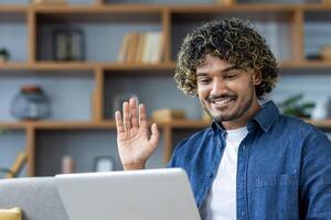 Man at home on sofa using laptop for call, young student waving happily at online conference meeting photo