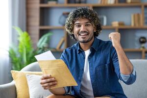 Winning good news, man with mail envelope looking joyfully at camera, sitting in living room at home, received notification message by mail about good results photo