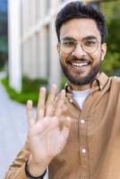 Bearded man with glasses and brown shirt smiling and waving while participating in call. Outdoor background with greenery and modern architecture. Concept of communication, positivity photo