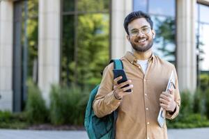 Happy Indian student with phone and backpack standing outside campus building photo