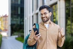 Indian student smiling while reading smartphone with backpack outside university campus photo