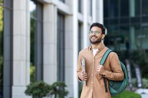 Student walking outside university campus with books in hand. Young man with backpack in casual clothes, happy and smiling. photo