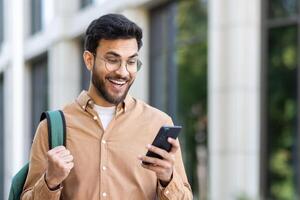 Man outside university campus with phone in hand celebrating successful exam results, student with backpack holding hand up in triumph gesture, received online notification on smartphone. photo