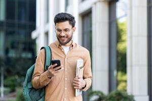 Young student with backpack on shoulder using app on smartphone. Man walking outside university campus smiling with phone in hands. photo