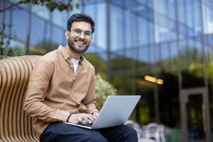 Smiling young man working on laptop outside modern building, enjoying remote work on sunny day photo