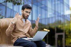 Frustrated man with laptop experiencing unexpected problem outdoors, expressing disbelief in urban setting on sunny day photo