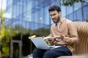 Young man sitting outdoors, looking frustrated while using laptop, expressing confusion, stress, and dissatisfaction during remote work on a sunny day photo