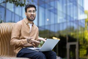 Confused man using laptop outdoors with modern glass building background, expressing frustration or misunderstanding during work, business or study session photo