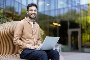 Smiling man sitting outdoors on a stylish bench while using a laptop, enjoying work or study in a modern urban environment with headphones photo
