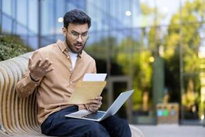 Shocked young man reading unexpected news in letter while sitting outside with laptop open, expressing surprise and confusion photo
