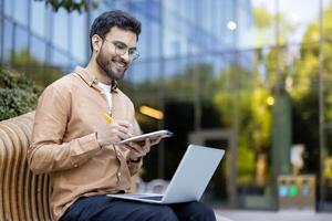 Smiling young professional takes notes on tablet while engaged in online learning with laptop in modern outdoor workspace photo