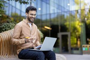 Smiling young man having call on laptop while sitting outside with earphones and notebook, enjoying remote work photo