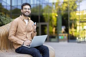 Happy man outdoors using laptop and holding asthma inhaler while taking a break from work in urban setting photo
