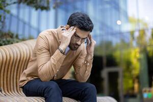 Stressed man sitting on bench holding head in hands while contemplating life and work challenges outside modern building photo