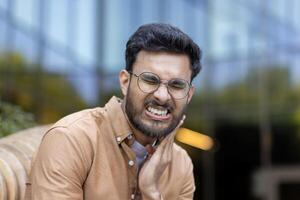 Man with glasses experiencing severe toothache outside office building, showing discomfort while holding cheek photo