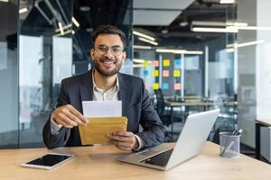 Confident businessman in modern office holding an envelope, smiling at the camera, with a laptop and tablet on the desk. Vibrant workspace with colorful notes in the background. photo