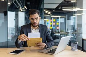 Serious man in a modern office holding a postal envelope, reading an important message. He is focused and appears thoughtful, surrounded by technology and office materials. photo