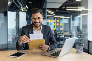 Business professional opening envelope while sitting at desk in modern office with laptop, tablet, and paperwork, looking pleased with correspondence received during workday photo