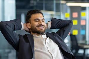 A well done man rests with his hands behind his head, dreams and thinks about the future results of his achievement. A businessman at his workplace inside the office works close-up in a business suit photo
