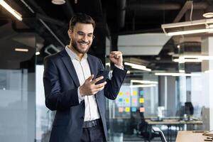 A businessman in a suit celebrates success in a modern office while looking at his smartphone, showcasing excitement and achievement in a professional setting. Bright office lights emphasize his joy. photo