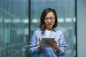 Serious and confident office worker using tablet computer. Asian businesswoman using online financial app, reading thoughtfully while standing near window inside office. photo