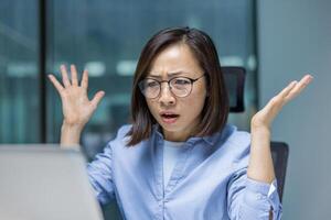 An Asian woman stares in disbelief at her computer screen in the office, experiencing frustration with an error or unexpected outcome. photo