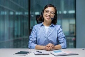 Webcam view, female support worker with headset smiling and looking at camera. Woman at workplace inside office, consulting and helping clients remotely online. photo