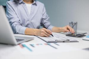A person diligently works on financial documents at a desk, using a calculator and laptop. The scene focuses on work and accounting activities photo