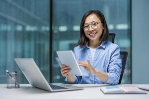 Asian businesswoman smiles at the camera while working at her desk with a laptop and tablet in a modern office setting. photo